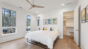 A modern bedroom with a large bed, a ceiling fan, and a view of trees through large windows. - Plutus' Playhouse (Palmetto)