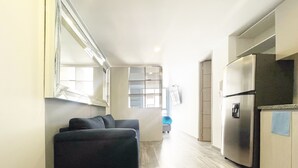 A narrow hallway with a dark sofa, a kitchen area with a stainless steel refrigerator, and a window with blinds.