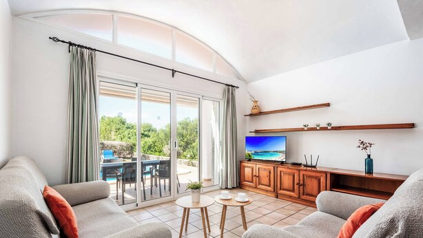A modern living room with a sofa, two small tables, a TV, and a view of a pool and greenery through large sliding glass doors.
