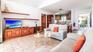 A spacious living area with a large flat-screen TV, wooden cabinets, a grey sofa, and a small dining table.