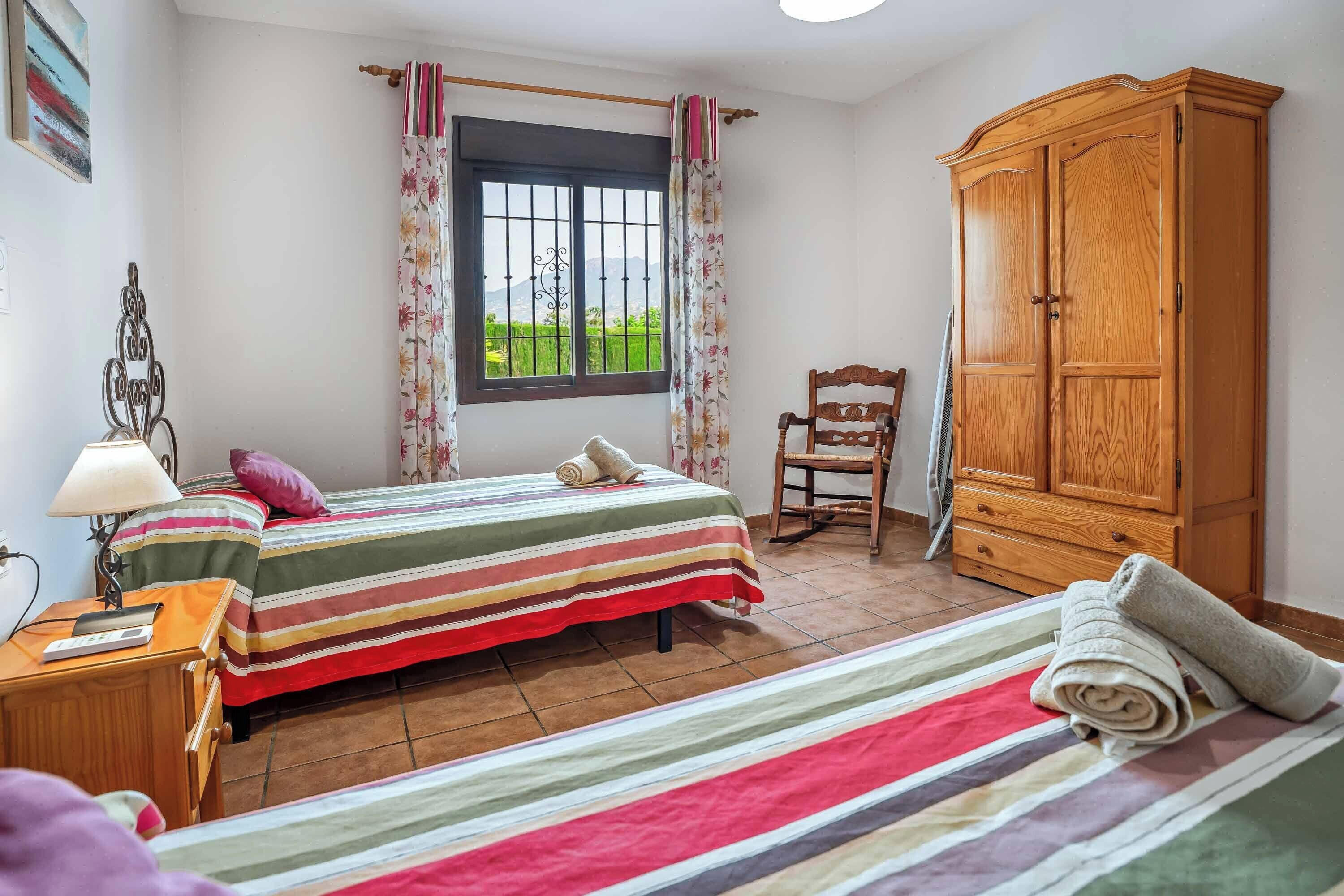 Une chambre avec deux lits, une armoire en bois, une chaise, une fenêtre avec des rideaux et une lampe sur une table de chevet.