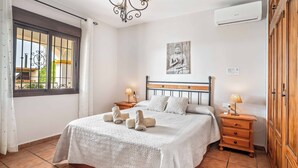A neatly made bed with a patterned bedspread and two pillows, a bedside table with a lamp, and a large window with bars. - Villa Los Sanchez - Two Bedroom Villa, Sleeps 4 (Frigiliana)
