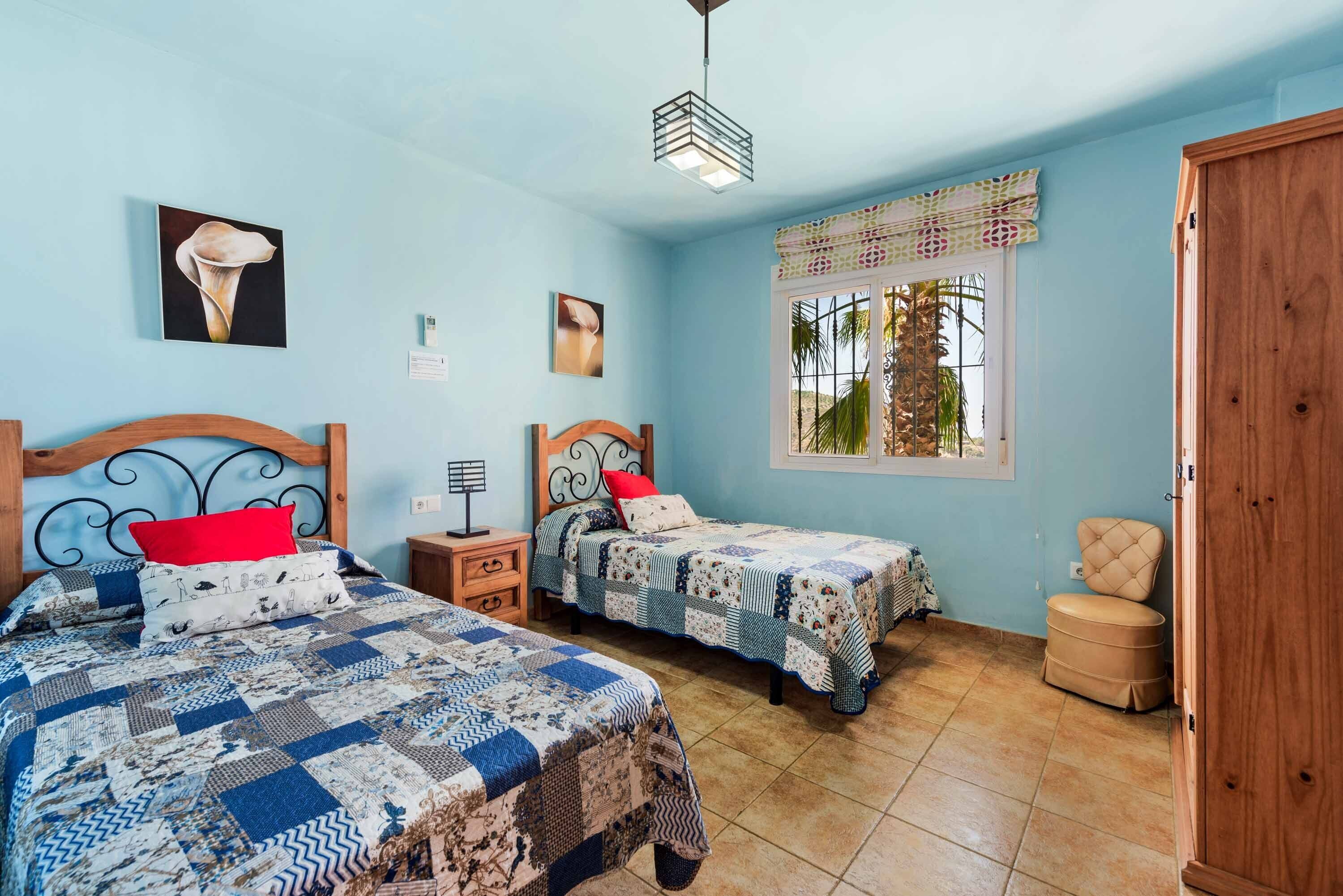 A bedroom with two beds, a wooden wardrobe, a window with a view of palm trees, and a ceiling light fixture.