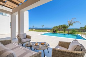 A poolside area with wicker furniture, a table with drinks, and a view of the sea.