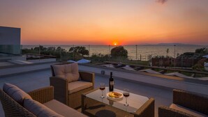 Vista sul tramonto da una terrazza panoramica con mobili in vimini, un tavolo in vetro e due bicchieri di vino rosso.