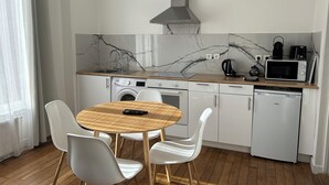 Cocina compacta con lavamanos, microondas, hervidor y lavadora. En primer plano, una mesa redonda de madera con sillas.