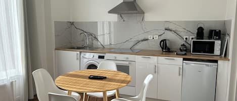 A compact kitchen with a sink, microwave, kettle, and a washing machine. There is a round wooden table with chairs in the foreground.