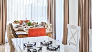 Un espace repas avec une longue table dressée pour le repas, entourée de chaises, et de grandes fenêtres ornées de rideaux.
