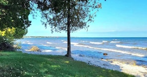 A beach with waves, a tree, and a grassy area. - Waterfront! New Construction The Resting Place (Sturgeon Bay)