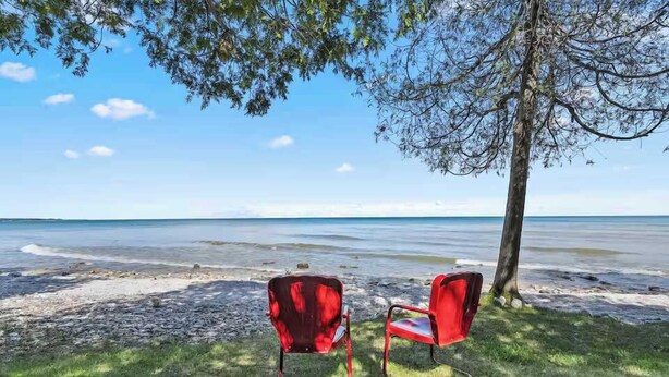 Two red chairs on a grassy area by a beach with a tree and a clear sky. - Waterfront! New Construction The Resting Place (Sturgeon Bay)
