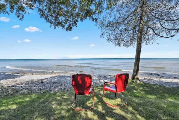 Two red chairs on a grassy area by a beach with a tree and a clear sky. - Waterfront! New Construction The Resting Place (Sturgeon Bay)