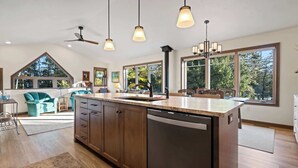 A modern kitchen with a large island, wooden cabinets, and a dishwasher. There are pendant lights, a ceiling fan, and large windows offering a view of the outdoors. - Waterfront! New Construction The Resting Place (Sturgeon Bay)