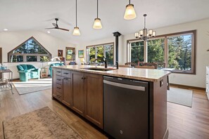 Une cuisine moderne dotée d’un grand îlot central, de meubles en bois et d’un lave-vaisselle. On y trouve des luminaires suspendus, un ventilateur de plafond et de grandes fenêtres offrant une vue sur l’extérieur.