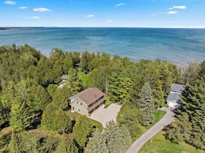 An aerial view of a property with a house surrounded by dense forest, a clear view of the ocean, and a paved driveway. - Waterfront! New Construction The Resting Place (Sturgeon Bay)