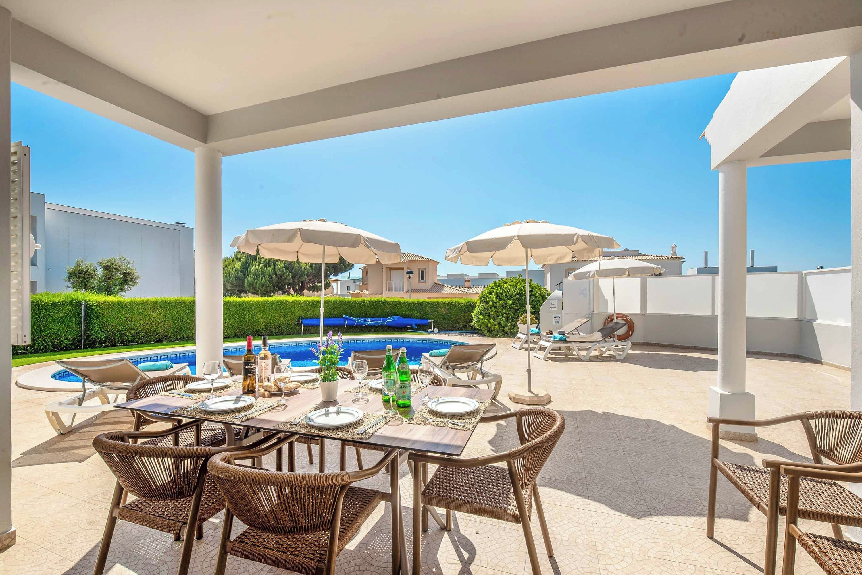 Un patio exterior con piscina, mesa de comedor, sillas, sombrillas y sillones de descanso.