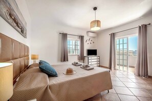 A hotel room with a bed, a television, a window with curtains, a wall-mounted air conditioner, and a balcony with a view.