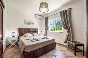 Ein Schlafzimmer mit einem großen Bett, Nachttischen, einer Bank, einer Hängelampe und einem Fenster mit Vorhängen.