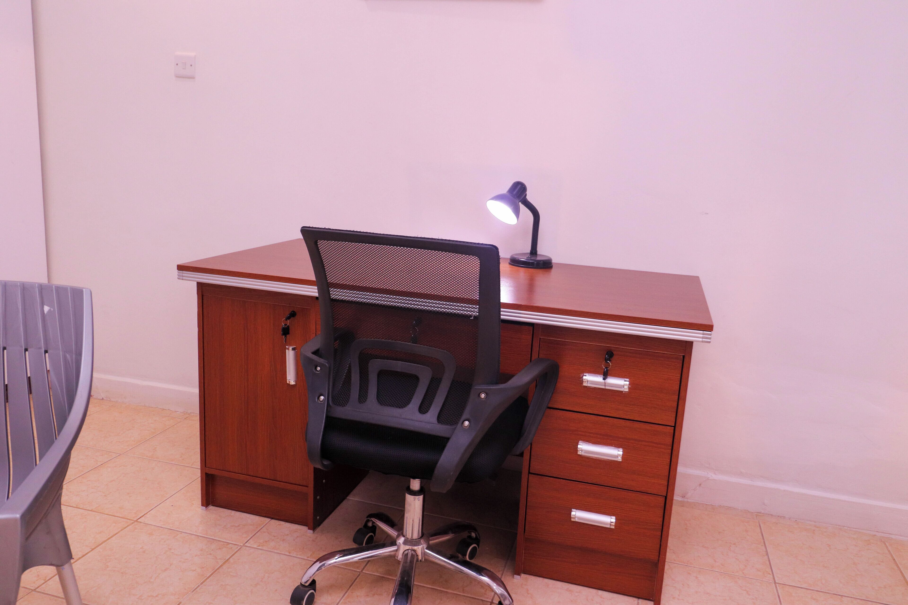 A wooden desk with a mesh chair, a desk lamp, and a grey chair.