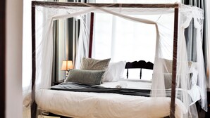 A four-poster canopy bed with a white bedspread and pillows, a bedside table with a lamp, and a view of the city through large windows.