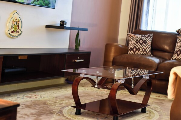 A living room with a glass-top coffee table, dark wooden entertainment unit, brown leather sofa, patterned carpet, and a wall-mounted television.
