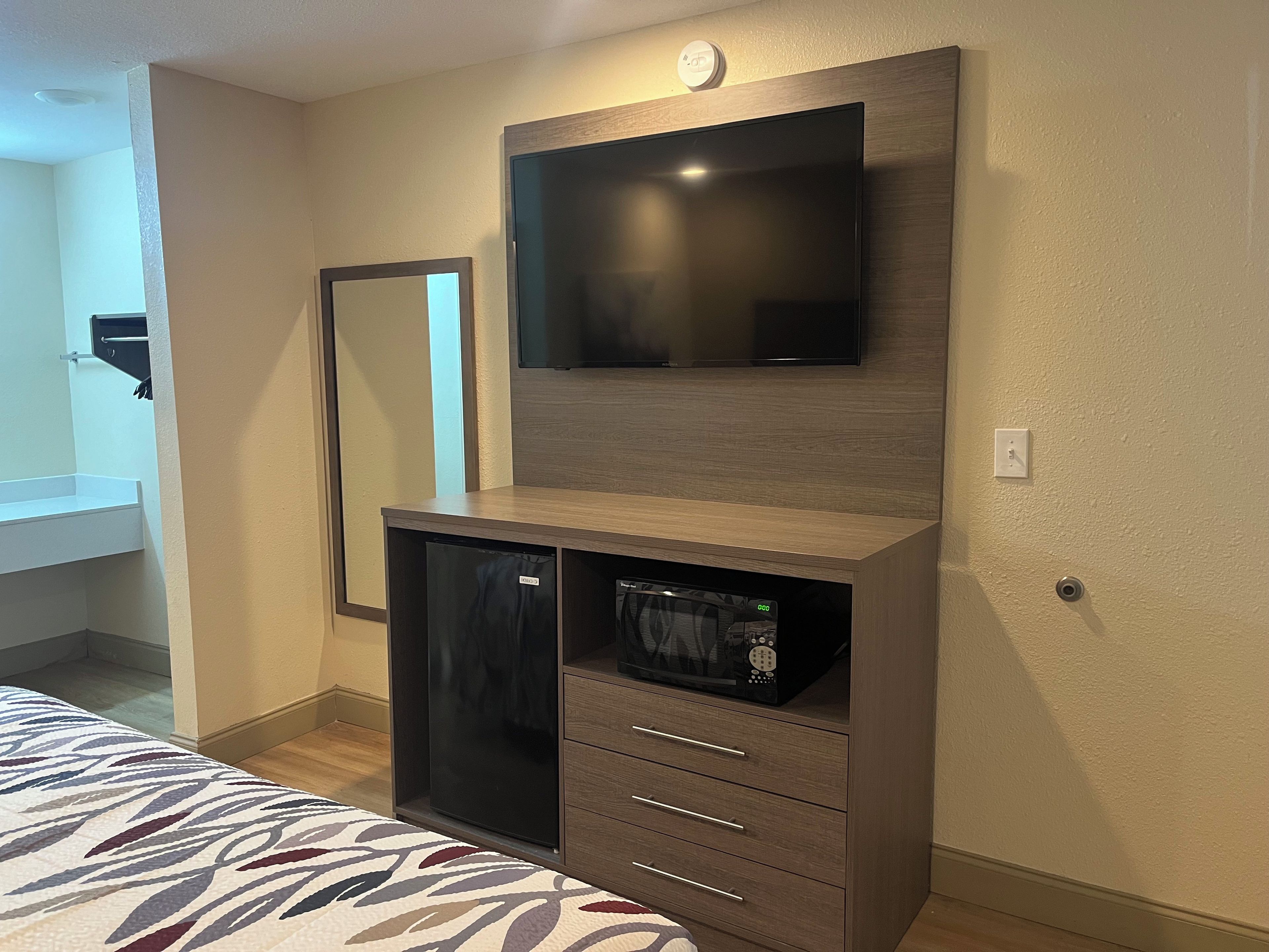 A compact room with a built-in TV, a mirror, a small refrigerator, and a bed with a patterned bedspread.
