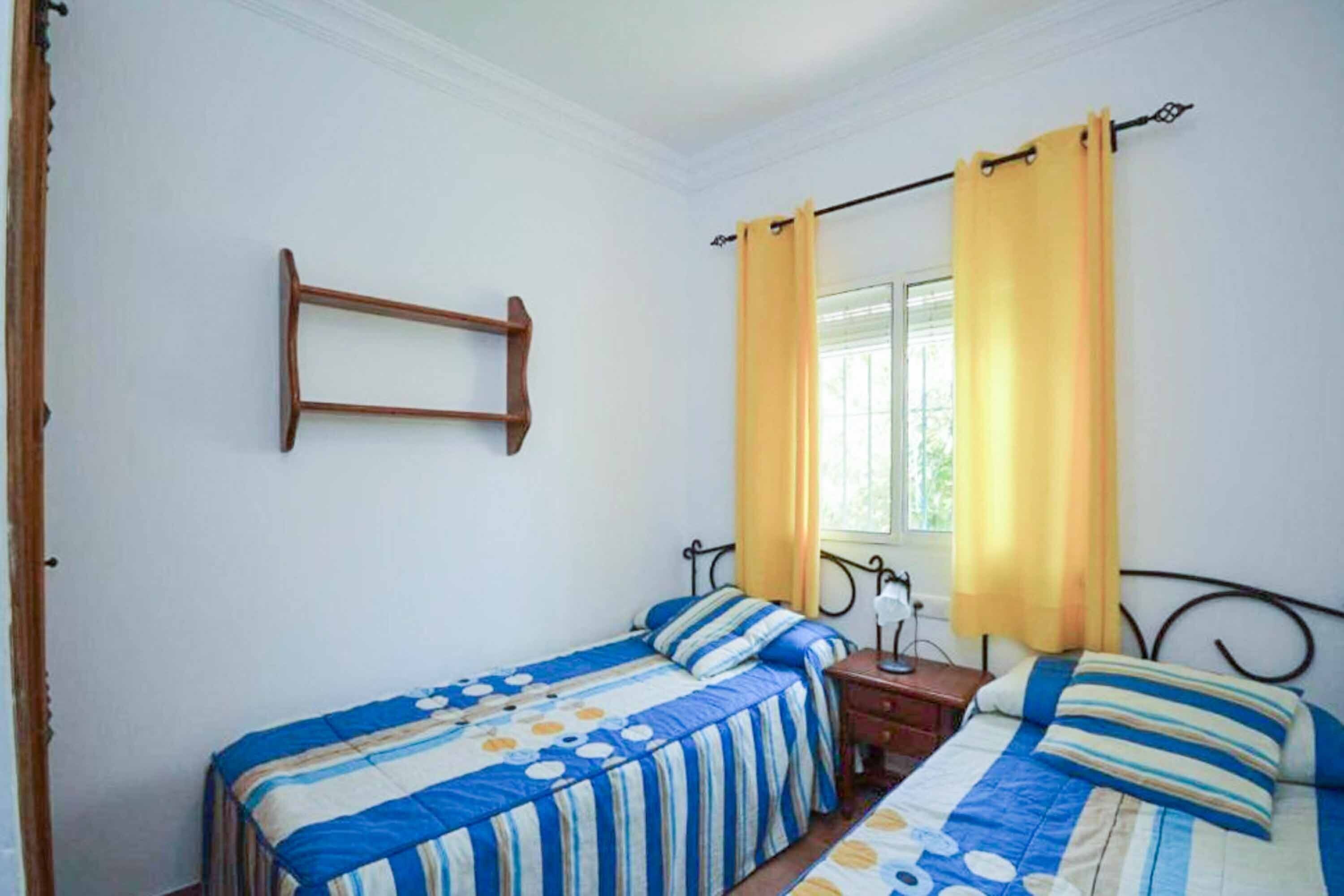 A bedroom with two beds, a window with yellow curtains, and a wooden shelf.