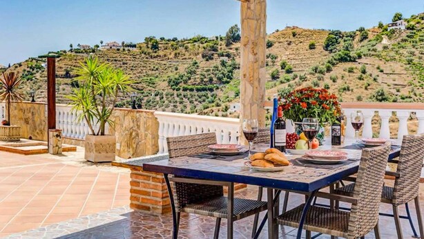 Un área de comedor al aire libre con una mesa dispuesta para dos personas, rodeada de sillas de mimbre y vistas a un paisaje montañoso.