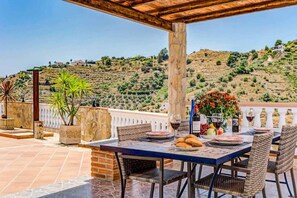 Un área de comedor al aire libre con una mesa dispuesta para dos personas, rodeada de sillas de mimbre y vistas a un paisaje montañoso.