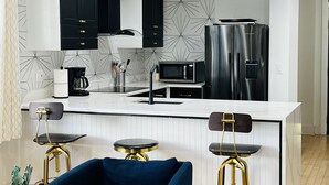 Une cuisine moderne avec des meubles noirs, un plan de travail blanc, un réfrigérateur en acier inoxydable, un four à micro-ondes et deux tabourets de bar aux pieds dorés.