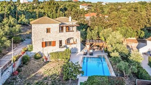 Una villa de piedra con piscina, rodeada de vegetación y otras viviendas.