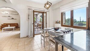 Una cocina moderna con una isla central, una mesa de comedor y sillas. En la zona de estar contigua hay un sofá, y se ve un patio por una puerta abierta.