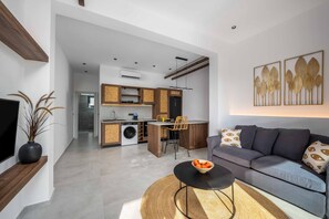 A modern living room with a grey sofa, a black coffee table, and a woven rug. There is a kitchen area with wooden cabinets and a dining space.
