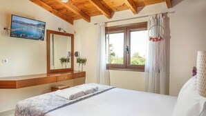 A bedroom with a bed, a wooden headboard, a large window with curtains, a vanity with a mirror, and a wall-mounted TV displaying a coastal scene. - Villa Portokalia Platanias (Platanias)