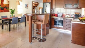 A modern kitchen with wooden cabinets, a central island with bar stools, a dining area with a table and chairs, and a staircase leading to an upper level. - Villa Portokalia Platanias (Platanias)