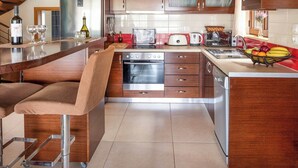 A modern kitchen with wooden cabinets, a built-in oven, a refrigerator, and a bar area with stools. - Villa Portokalia Platanias (Platanias)