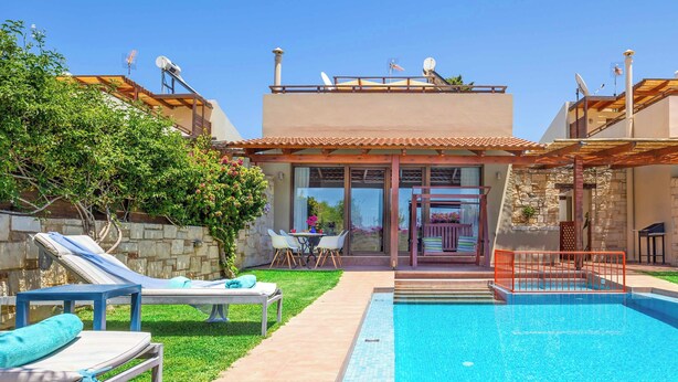 A swimming pool with lounge chairs, a stone wall, and a building with a tiled roof and satellite dishes. - Villa Portokalia Platanias (Platanias)