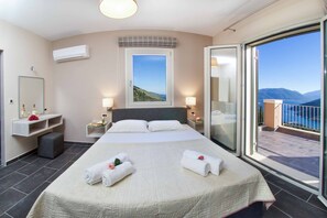 A hotel room with a large bed, a balcony with a view of the sea, and a window with a view of the hills.
