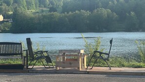 Zona con bancos junto al lago, con un banco de madera y dos sillas de metal, con vistas a un lago tranquilo y una colina boscosa al fondo.