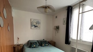 Un dormitorio con cama, una mesita de noche, un radiador, una ventana con cortinas, un cuadro colgado en la pared y una alfombra estampada.