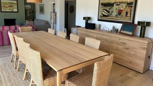 A dining area with a long wooden table and wicker chairs, a wooden cabinet with decorative items, and a painting of a boat on the wall. - Location Maison Carnac Plage (CARNAC PLAGE)