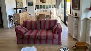 A spacious living area with a striped sofa, a dining table, and a kitchen in the background. - Location Maison Carnac Plage (CARNAC PLAGE)