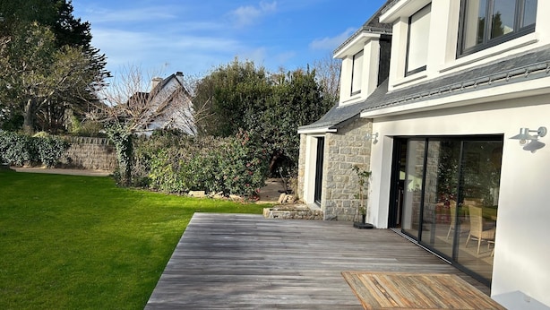 A modern house with a wooden deck, a glass door entrance, and a stone wall in the background. - Location Maison Carnac Plage (CARNAC PLAGE)