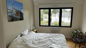 A bedroom with a large bed, a window with a view of buildings, a radiator, and a chair with a floral pattern. - Location Maison Carnac Plage (CARNAC PLAGE)