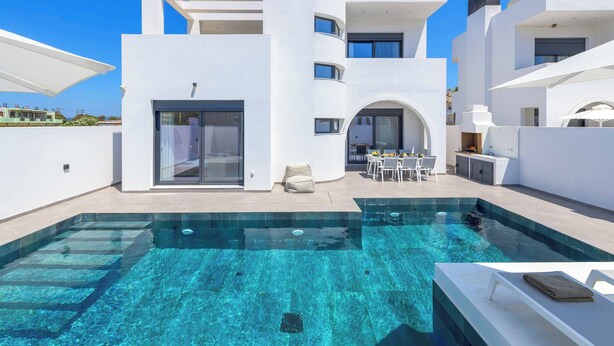 Un bâtiment moderne de couleur blanche, doté d’une piscine, d’un espace extérieur aménagé et surplombé d’un ciel bleu azur.