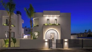 Un bâtiment moderne doté d’une entrée sécurisée par un portail, éclairé par un système d’éclairage d’ambiance et entouré de palmiers.