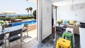A modern hotel room with a balcony overlooking a pool and the sea, a sofa, a dining table, and a small kitchen area. - Villa Cyan Paphos - Three Bedroom Villa, Sleeps 6 (Pafos)