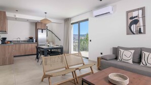 A modern apartment with a kitchen, dining area, and living room. It includes wicker furniture, a grey sofa, and a view of the city. - Massari Hills Villa 8 (Rhodes)
