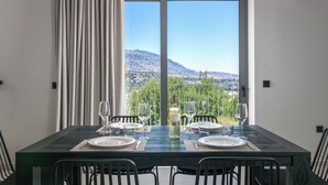 A dining table set for two with a bottle of wine, glasses, and plates, overlooking a mountainous landscape through a large window. - Massari Hills Villa 8 (Rhodes)