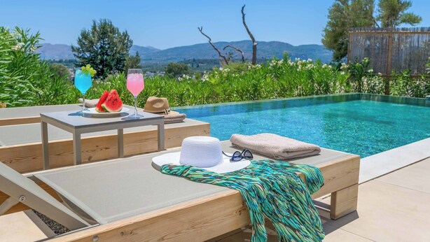 A poolside area with a wooden deck, lounge chairs, and a table with drinks and a hat. - Massari Hills Villa 8 (Rhodes)
