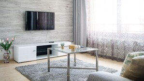 Una sala de estar moderna con un televisor de pantalla plana montado en una pared de madera, una mesa de centro de vidrio, un sofá blanco y un florero decorativo con flores.